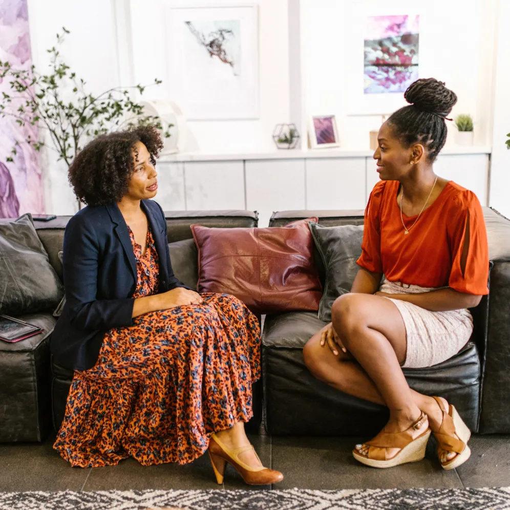 Two women sitting on a sofa, engaged in a discussion, with one woman gesturing while the other listens attentively